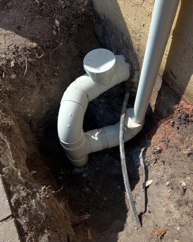 New white PVC bend with an inspection cap fitted in an open trench beside a downpipe